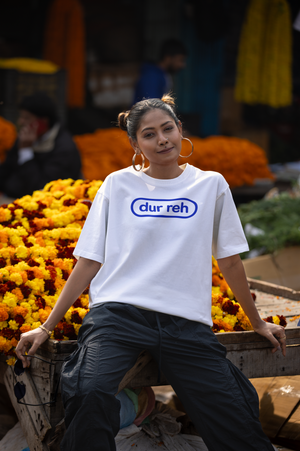 Woman wearing a white t-shirt with 'dur reh' text, standing amidst flowers.